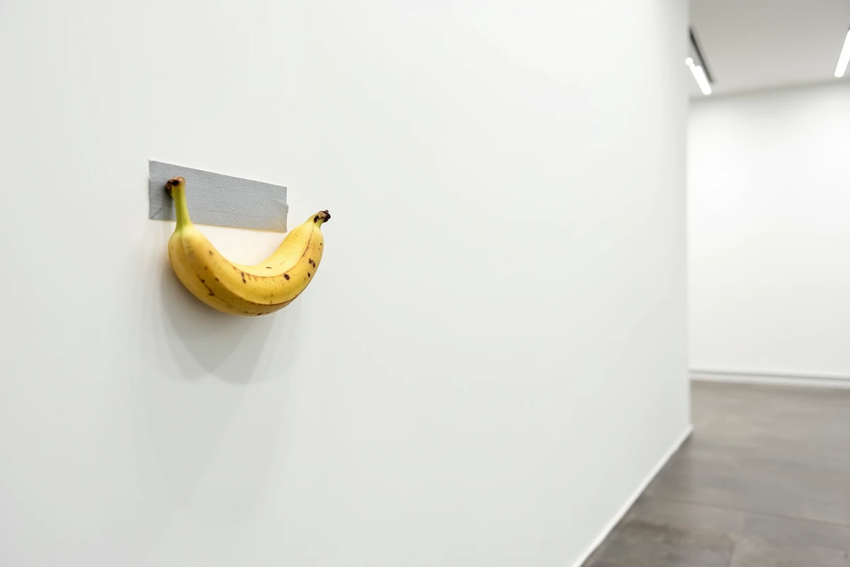 Une banane fixée avec du ruban adhésif argenté sur un mur blanc de galerie d'art contemporain, œuvre conceptuelle minimaliste.